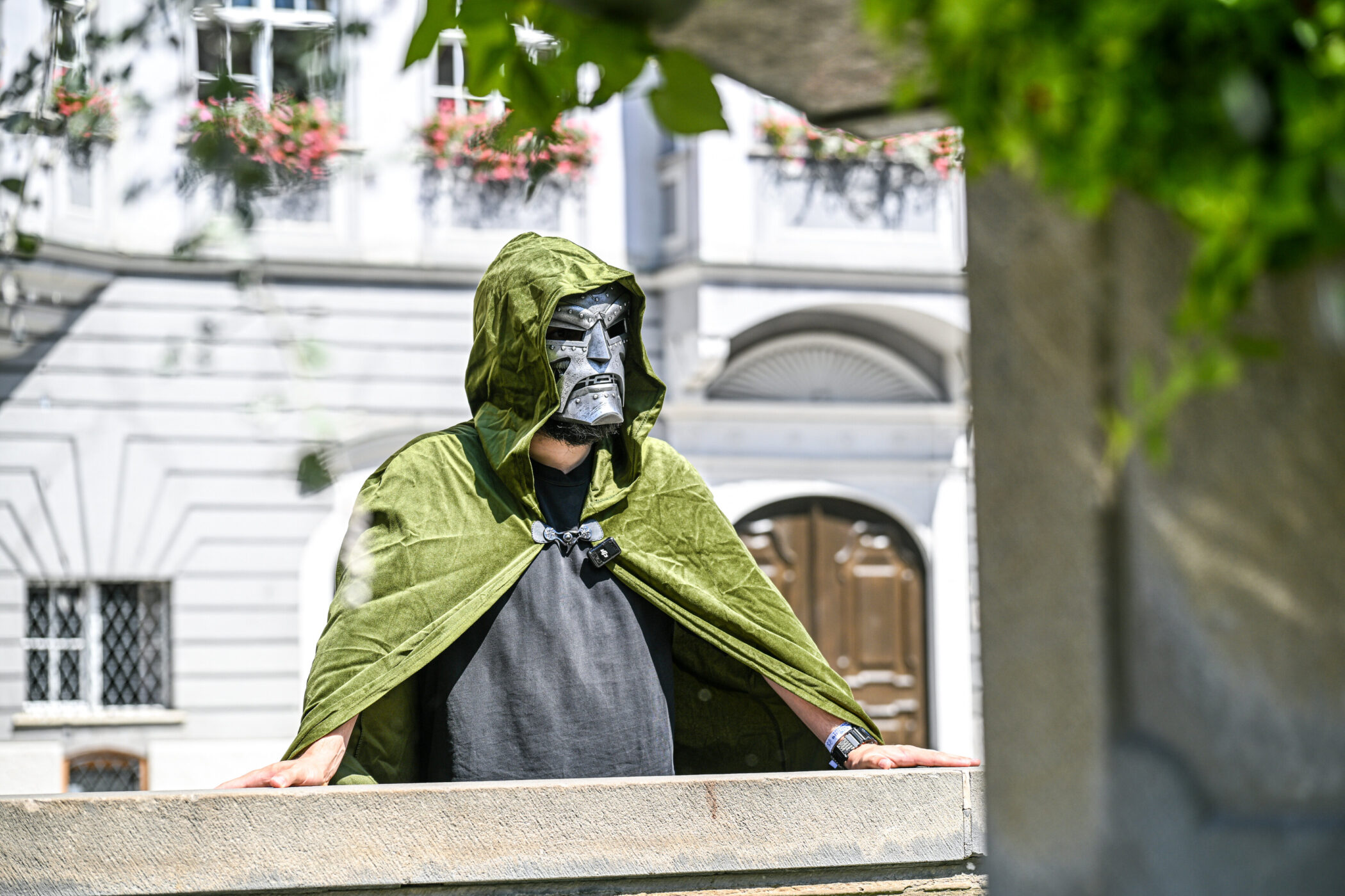 Person in grünem Umhang mit Maske, im Freien vor historischer Fassade.