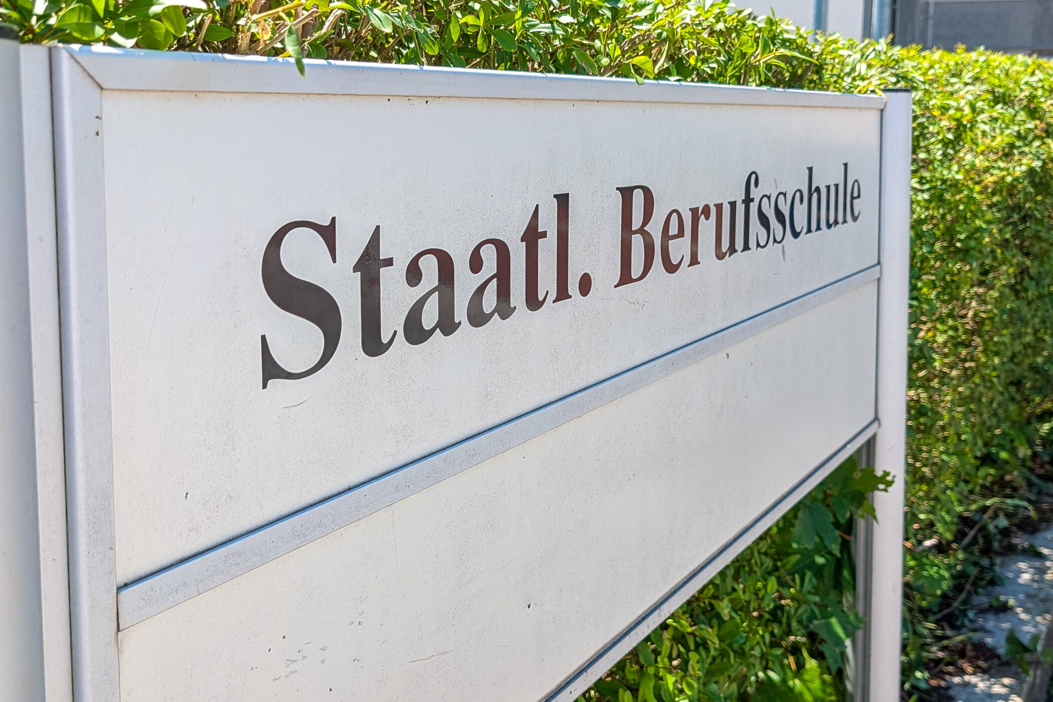 Schild mit der Aufschrift "Staatl. Berufsschule" vor einer Hecke.