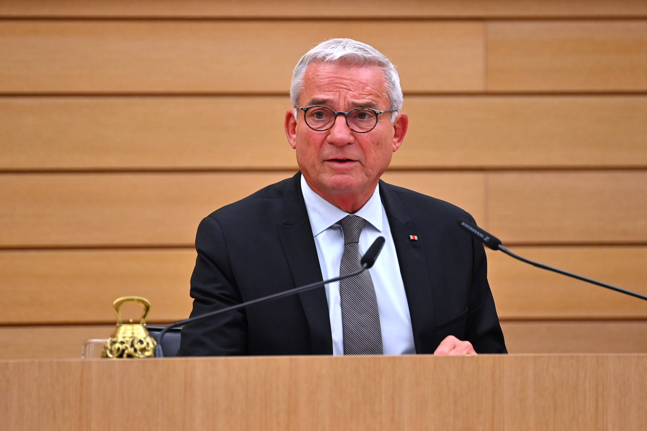 Älterer Mann im Anzug spricht an einem Pult mit Mikrofonen.