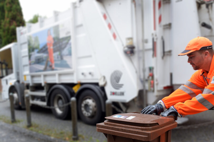Müllwerker schiebt eine braune Biotonne, im Hintergrund ein Müllwagen.