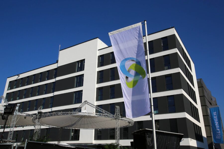 Modernes Gebäude mit Flagge, darauf blau-grünes Logo. Rechts ein Banner mit "Bilfinger".