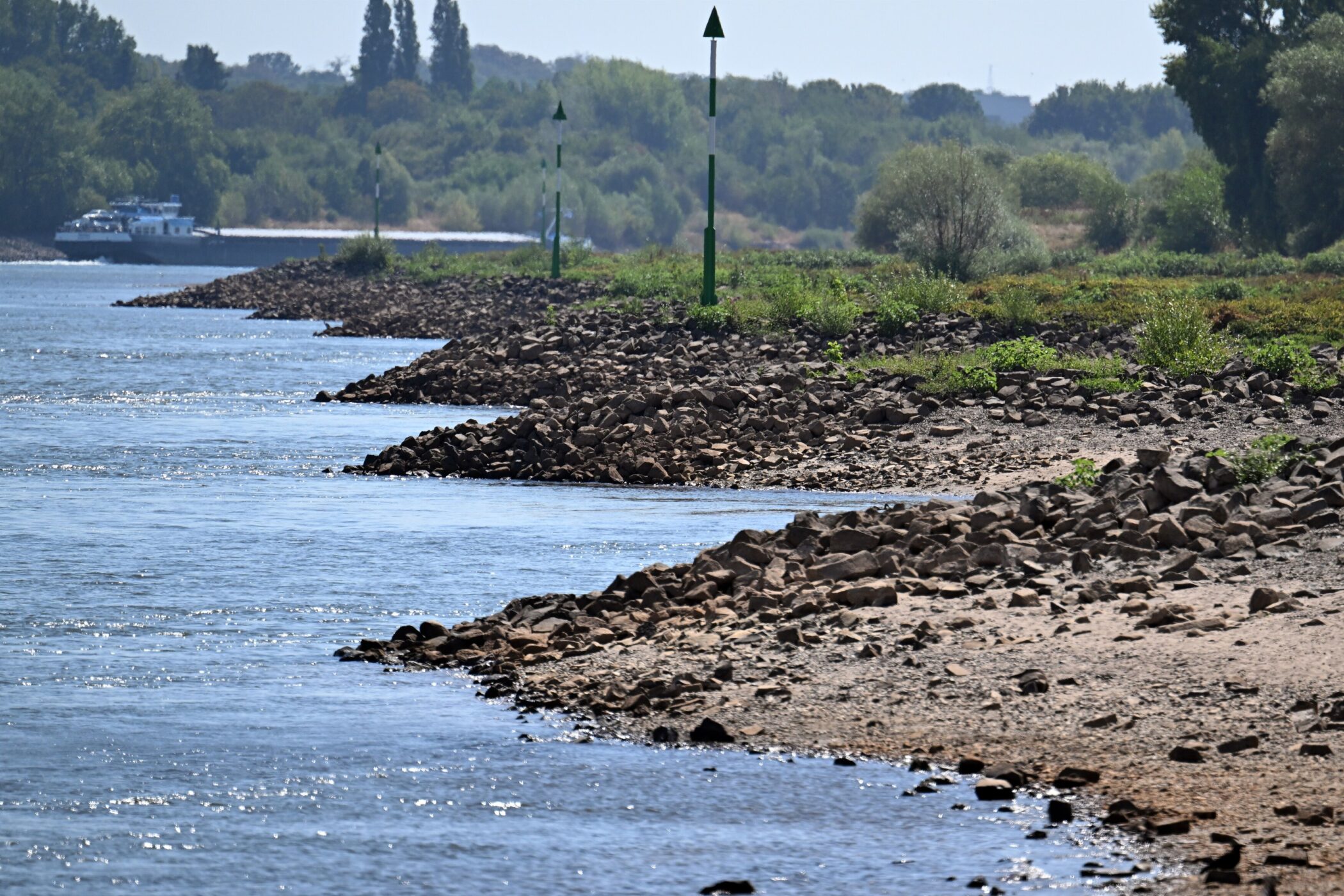 Strom, Ufer mit Steinen, grüne Markierungen, Bäume im Hintergrund, Boot in der Ferne.