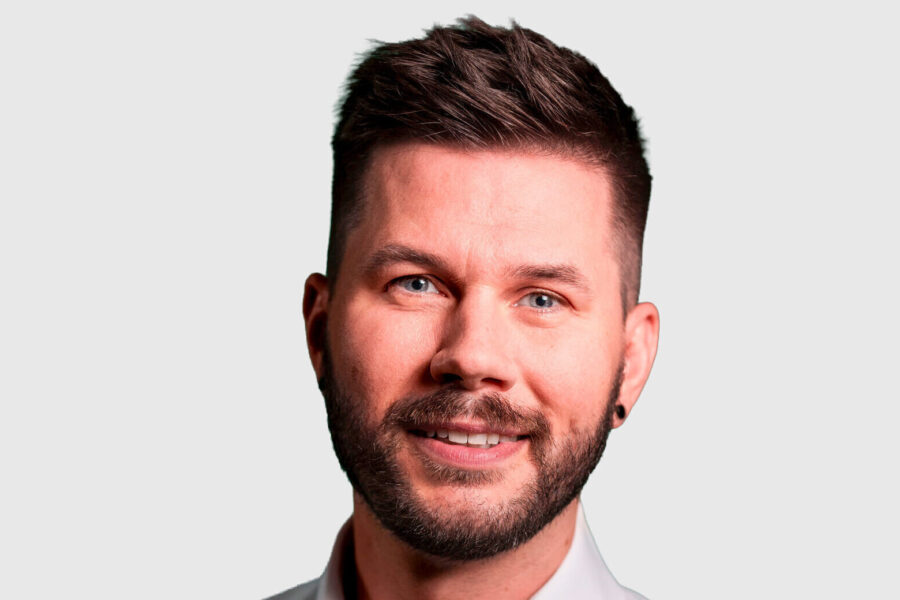 Ein Mann mit Bart und Ohrring lächelt vor einem neutralen Hintergrund.