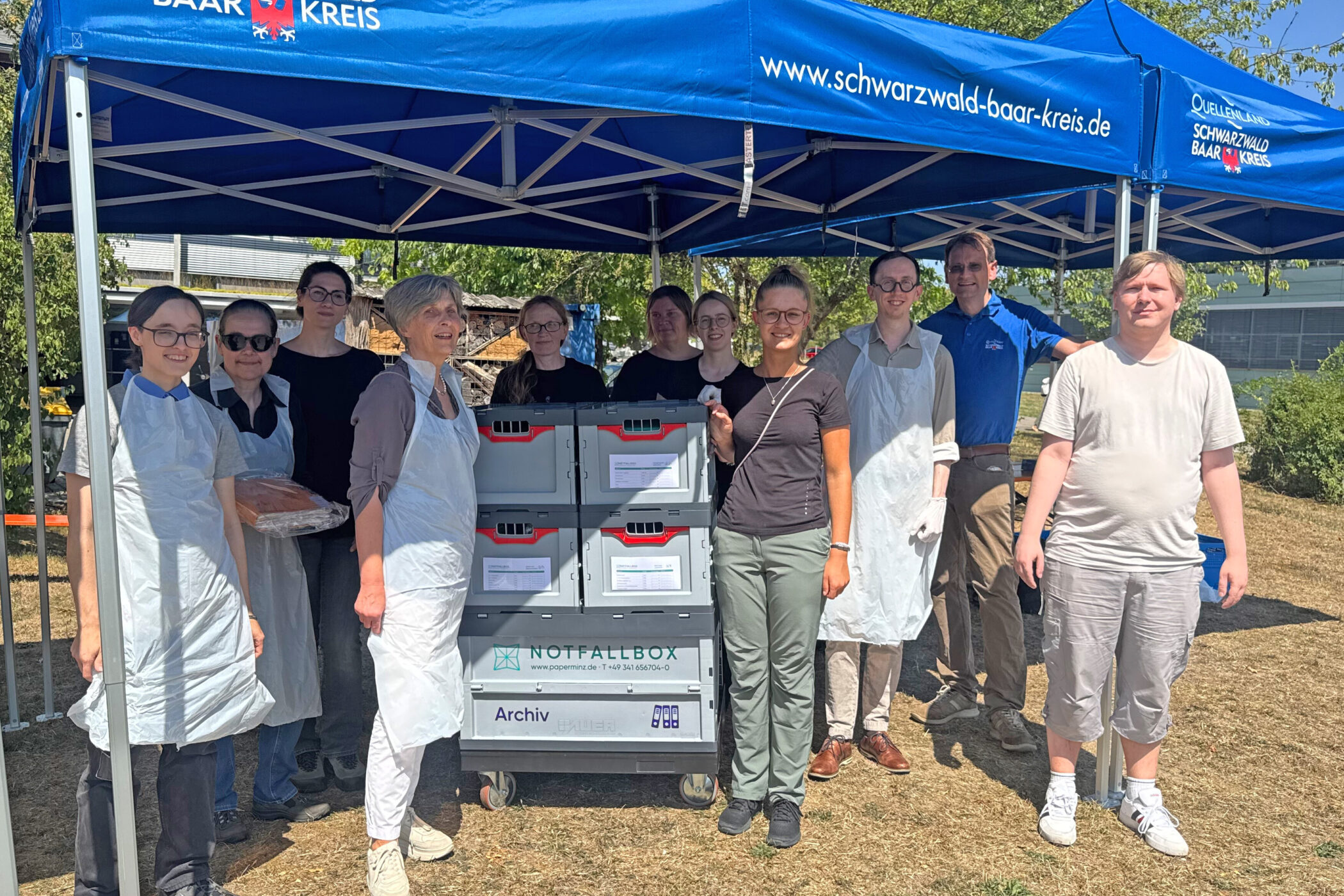 Gruppe von elf Personen unter blauem Pavillon, Notfallboxen im Vordergrund.