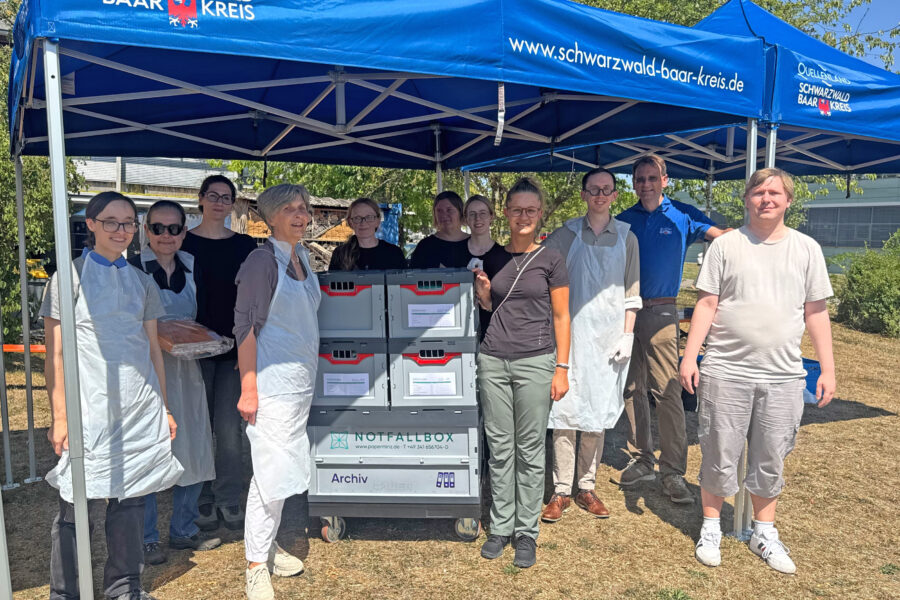 Gruppe von elf Personen unter blauem Pavillon, Notfallboxen im Vordergrund.
