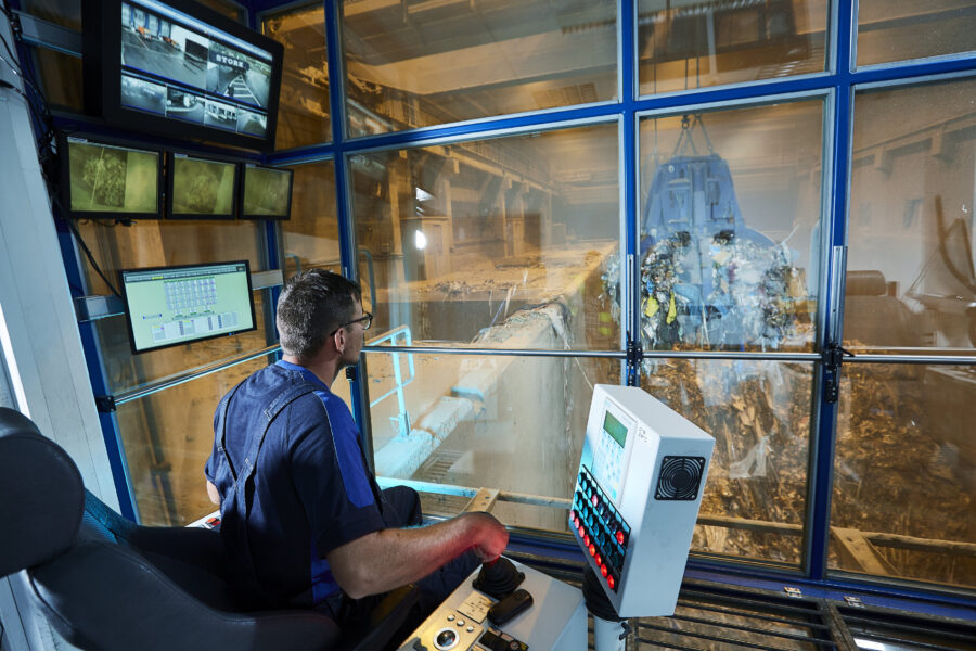 Mensch steuert Hebel und schaut durch Fenster, Monitore an Wand.