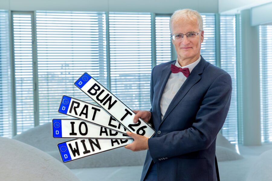 Person hält vier weiße Nummernschilder mit schwarzem Text „BUN T26“, „RAT ER“, „IO ADW“, „WIN ADW“.
