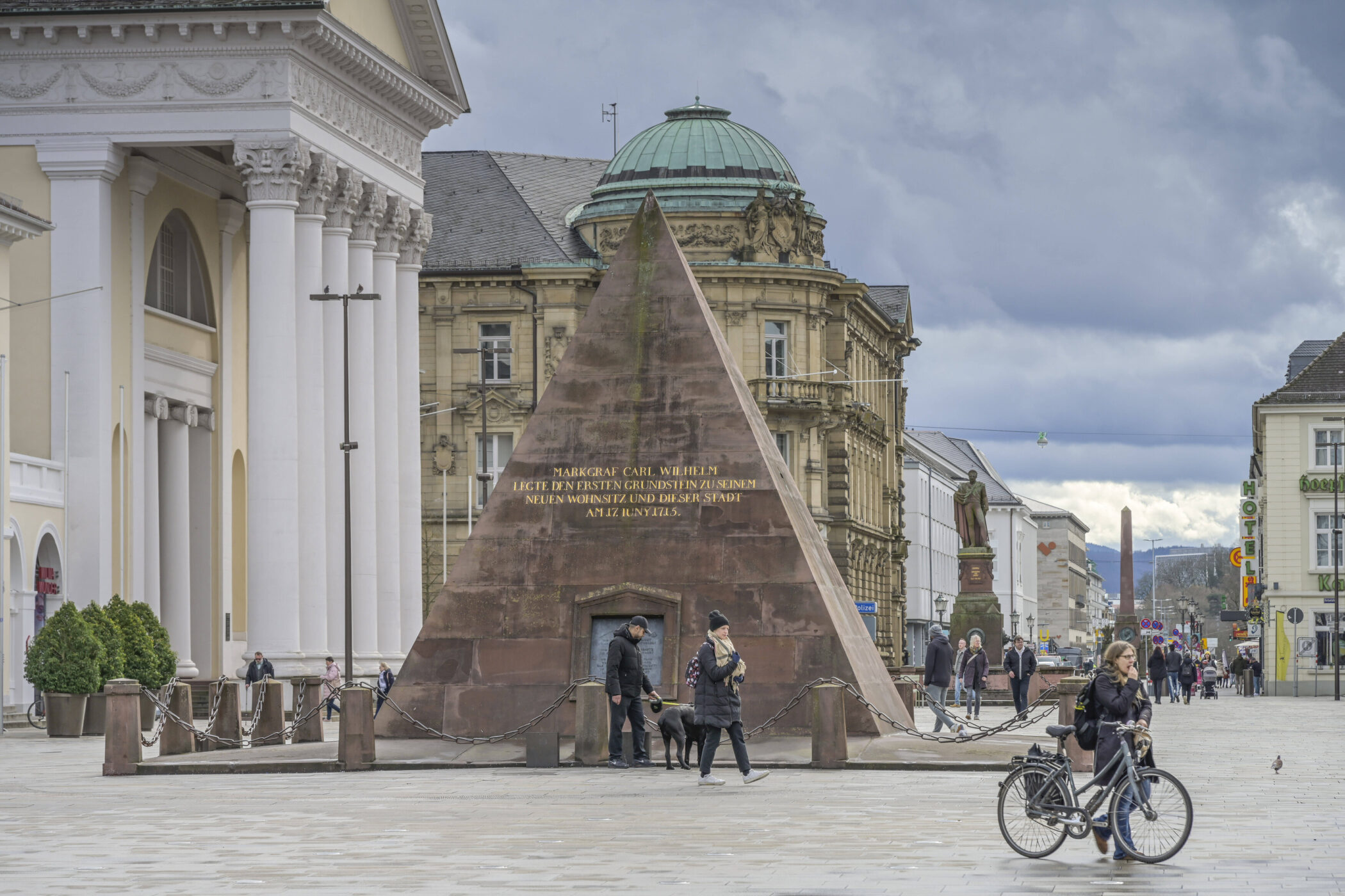 Steinpyramide mit Inschrift, Menschen gehen vorbei, Frau mit Fahrrad im Vordergrund.