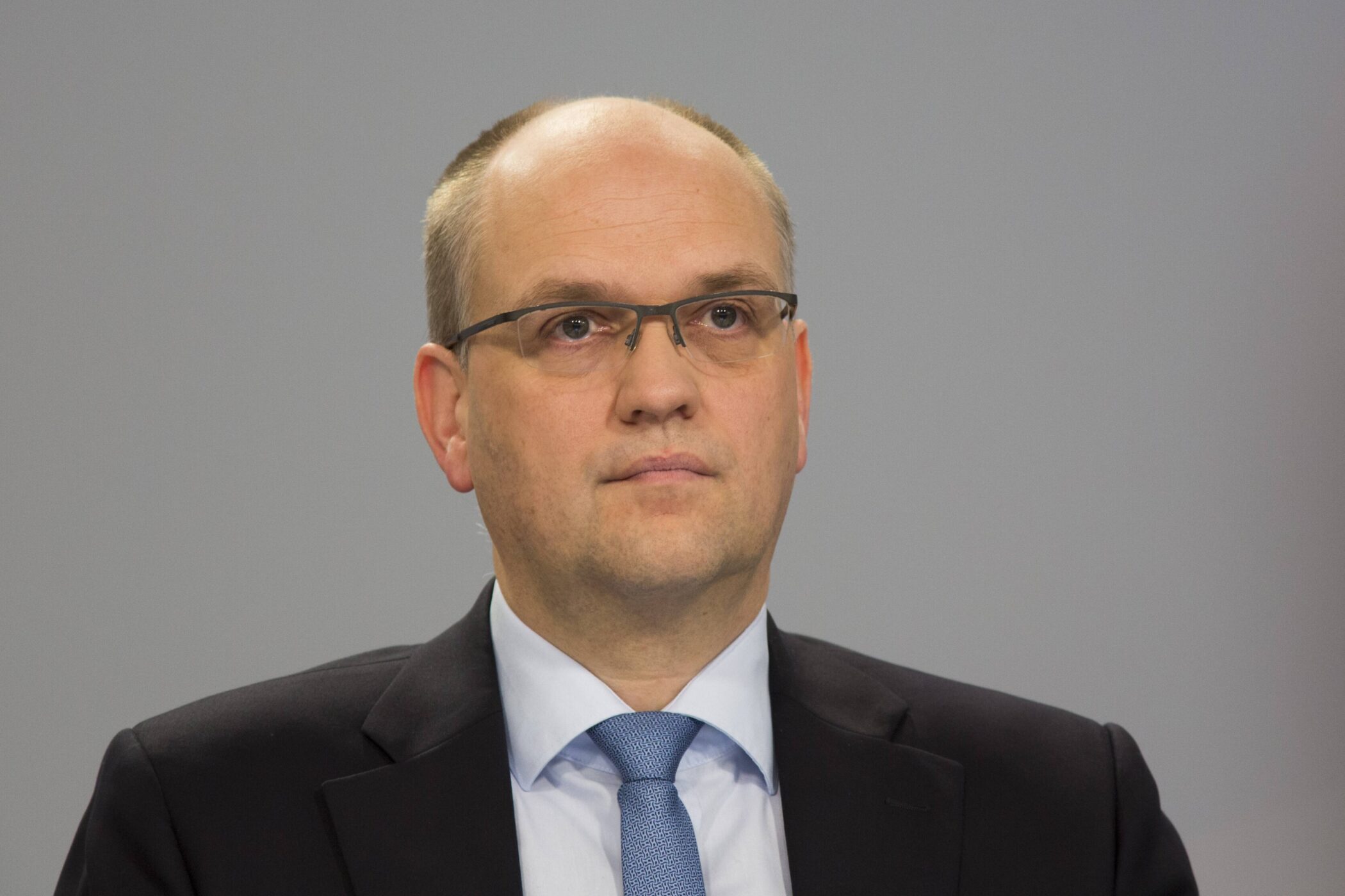 Ein Mann mit Brille und Anzug vor grauem Hintergrund.