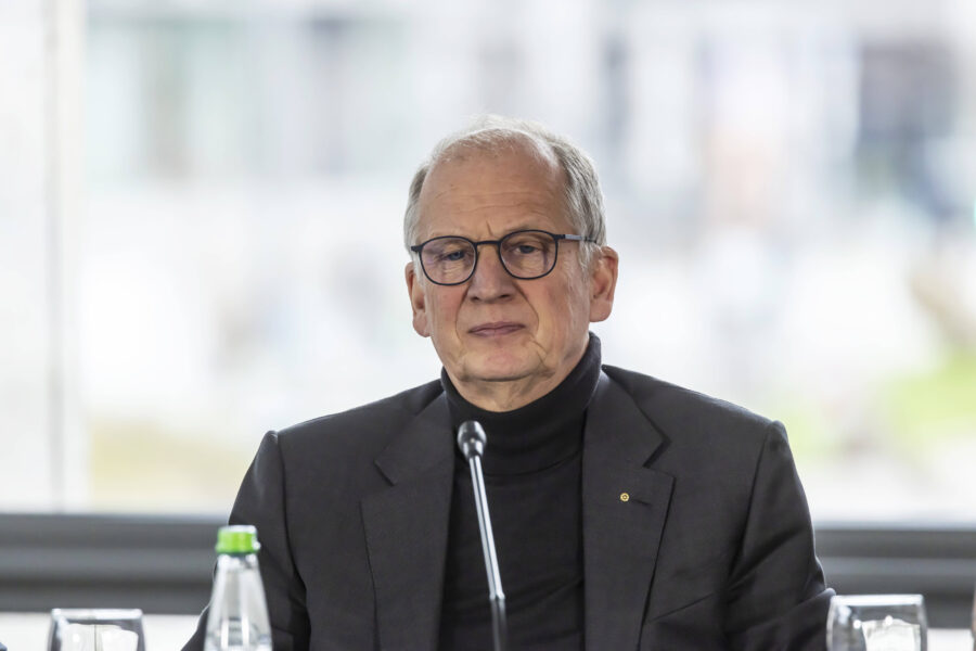 Eine Person mit Brille spricht in ein Mikrofon vor verschwommenem Hintergrund.