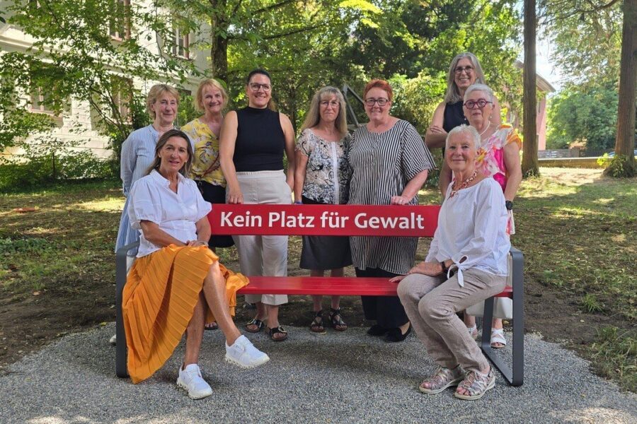 10 Personen, 2 sitzen auf Bank mit Text "Kein Platz für Gewalt", im Freien.