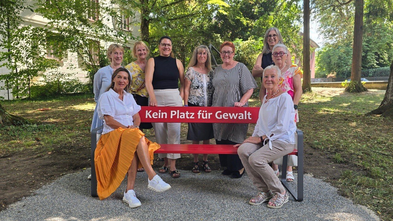 10 Personen, 2 sitzen auf Bank mit Text "Kein Platz für Gewalt", im Freien.