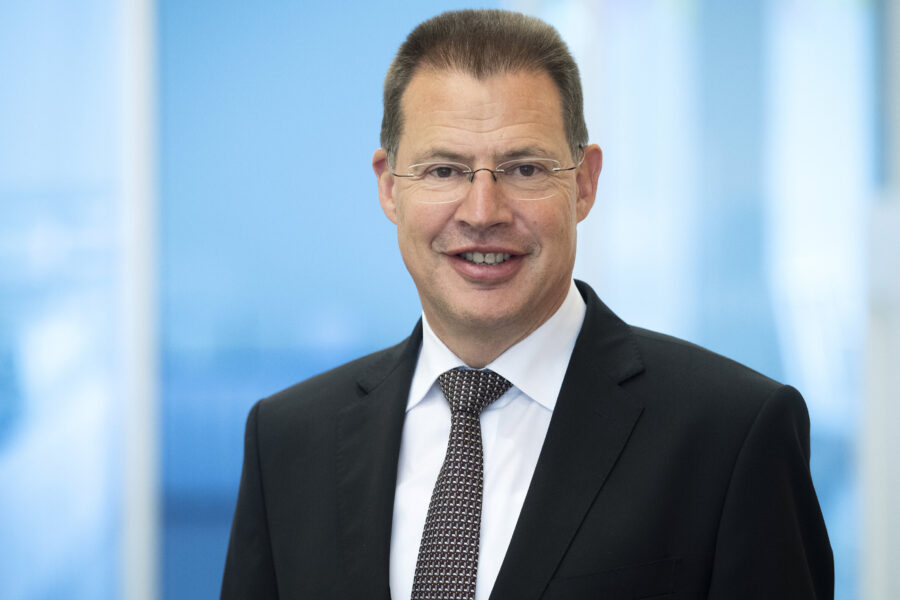 Mann mit Brille und Anzug lächelt vor unscharfem blauem Hintergrund.