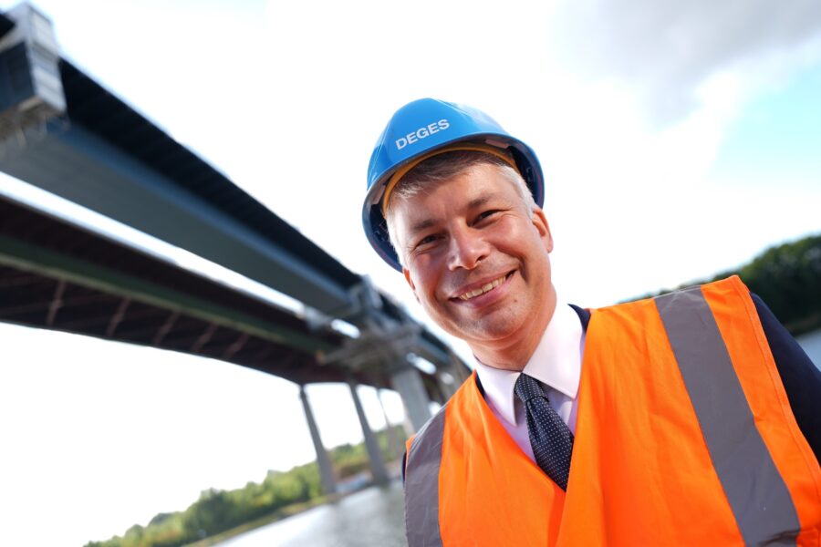 Person mit Blauhelm und Warnweste vor Brücke. Helmtext: "DEGES".