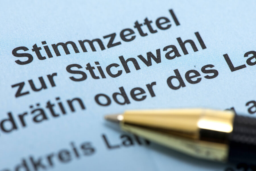 Text „Stimmzettel zur Stichwahl“ auf blauem Papier, Stift im Vordergrund.