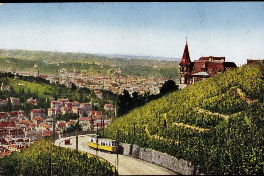 Gelbe Straßenbahn auf kurviger Straße, Häuser und Landschaft im Hintergrund.