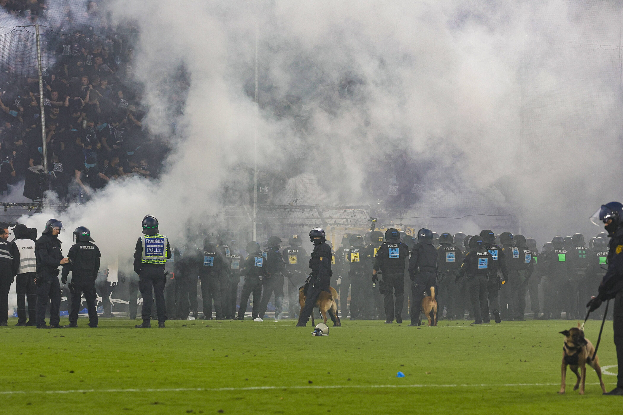 Polizisten und Hunde im Stadion, Rauch in der Luft, Menschenmenge im Hintergrund.