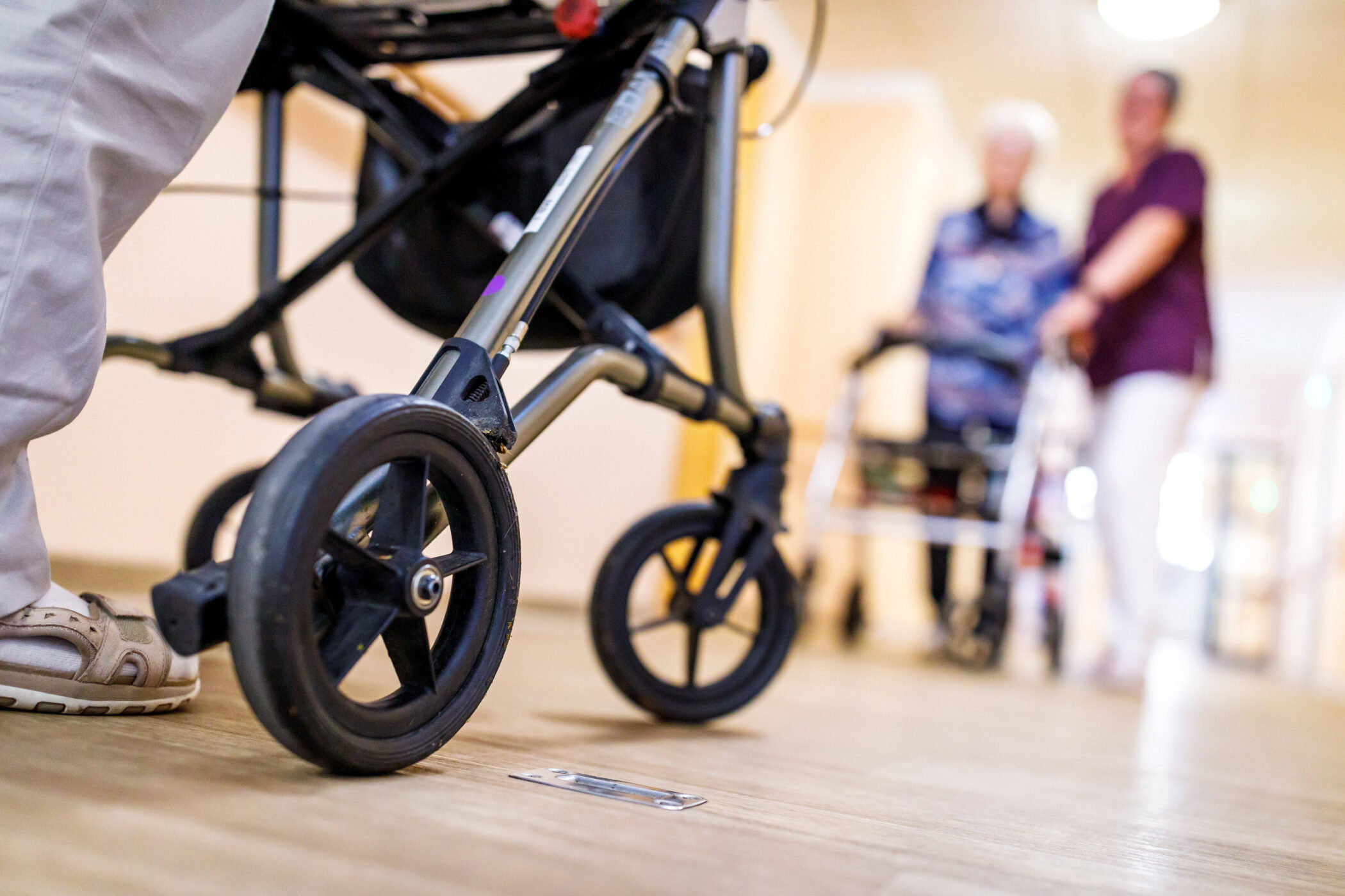 Rollator auf Holzboden, im Hintergrund zwei Menschen unscharf erkennbar.