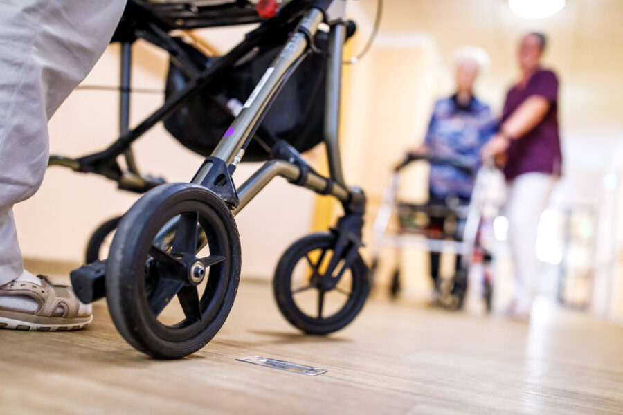 Rollator auf Holzboden, im Hintergrund zwei Menschen unscharf erkennbar.