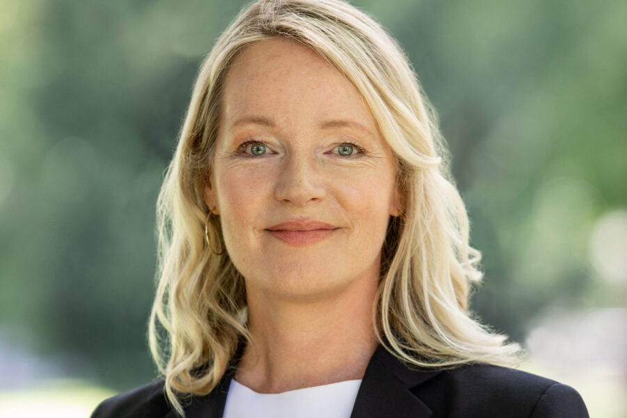 Frau mit blondem Haar in einem schwarzen Blazer vor grünem Hintergrund.