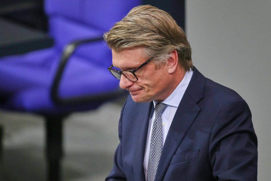 Mann im Anzug mit Brille, blauer Stuhl im Hintergrund.