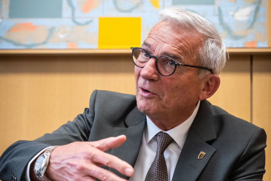 Älterer Mann mit Brille in Anzug gestikuliert vor abstraktem Kunstwerk.