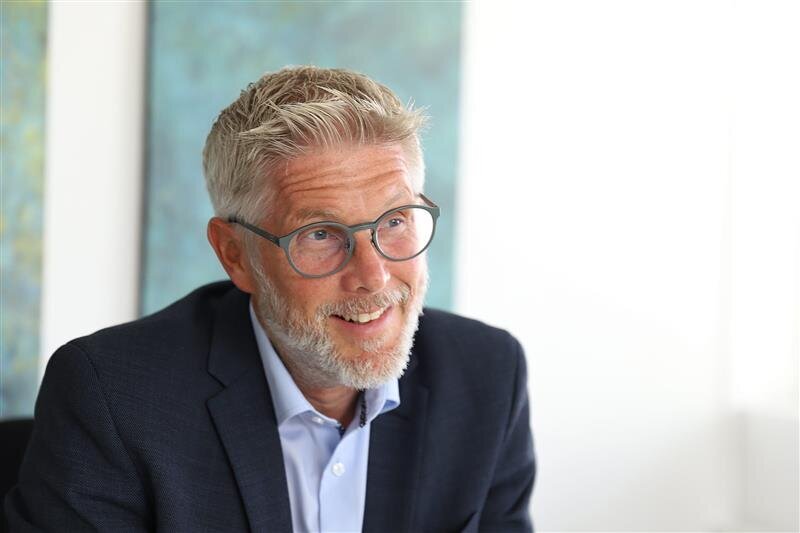 Mann mit Brille und Bart lächelt, trägt Anzug und Hemd, vor unscharfem Hintergrund.
