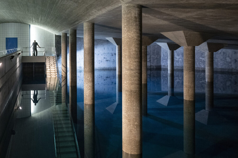 Unterirdischer Raum mit Wasser, Säulen und eine Person auf einer Treppe.