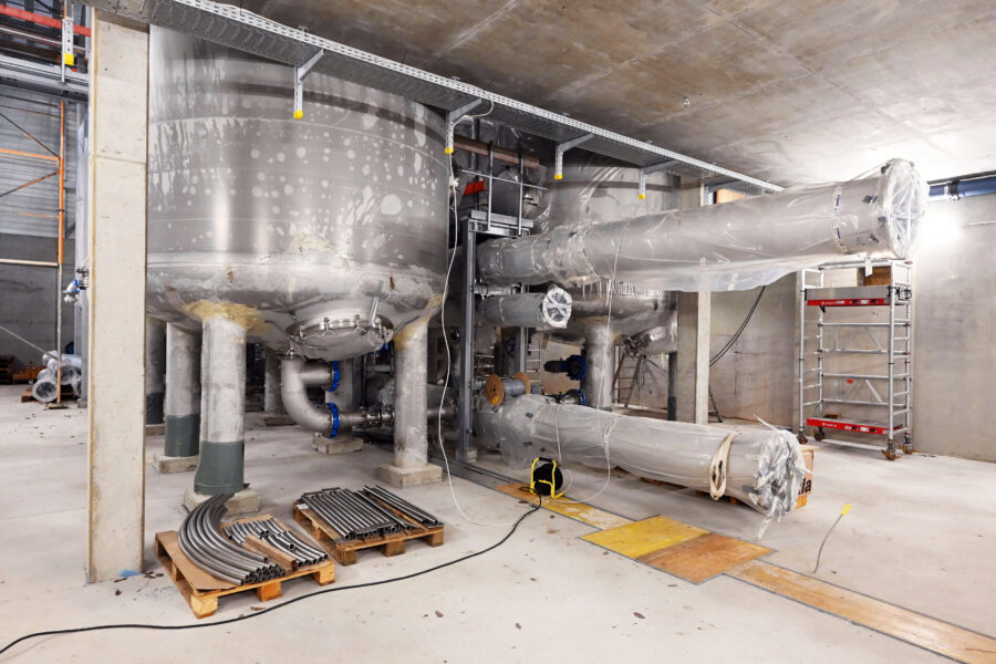 Große Metalltanks und Rohre in einem industriellen Raum, daneben Leitern und Paletten.