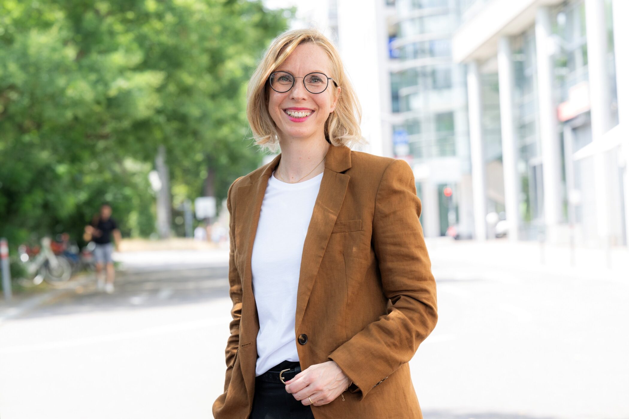 Person mit Brille und braunem Blazer draußen lächelnd.