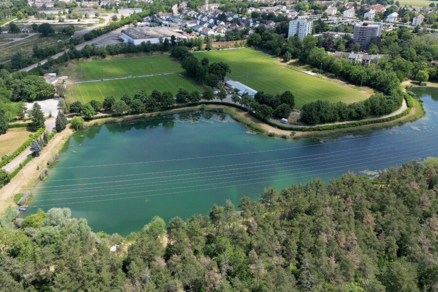 Wald, See und Sportplatz mit Bäumen umrandet, Stromleitungen über dem Wasser.