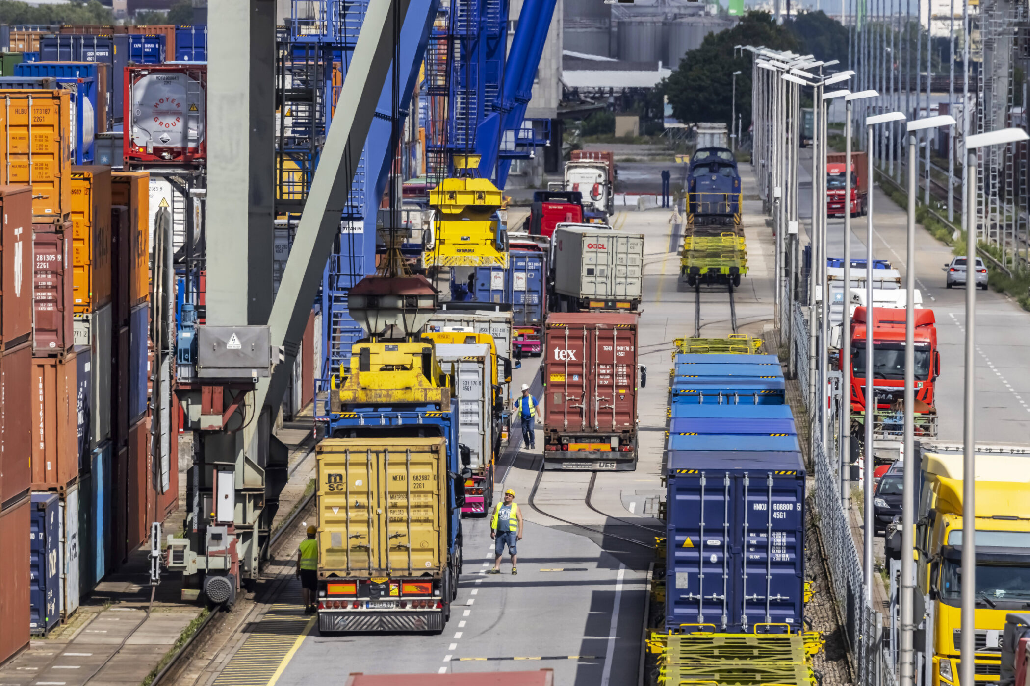 Hafen mit Containern, LKWs und Lokomotive. Zwei Personen arbeiten auf der Straße.