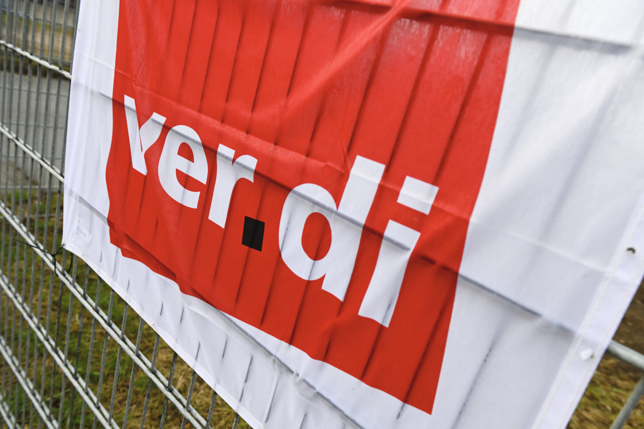 Rotes Banner mit weißem Text: "ver.di", an einem Metallzaun befestigt.