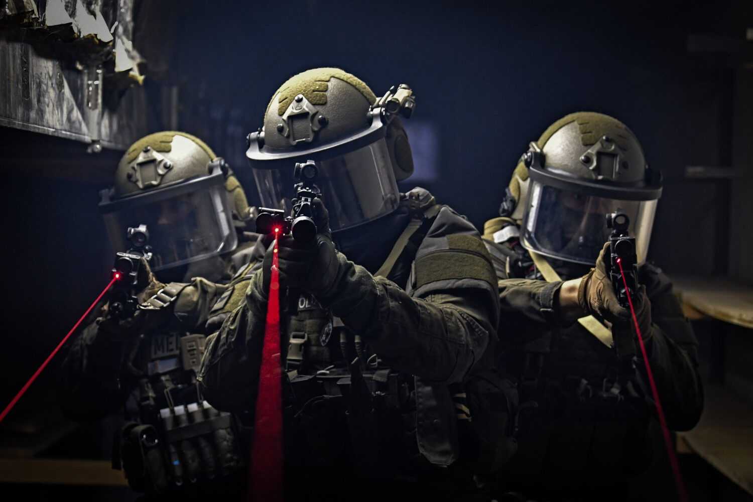 Drei uniformierte Personen mit Helmen, halten Gewehre mit roten Laserzielgeräten.