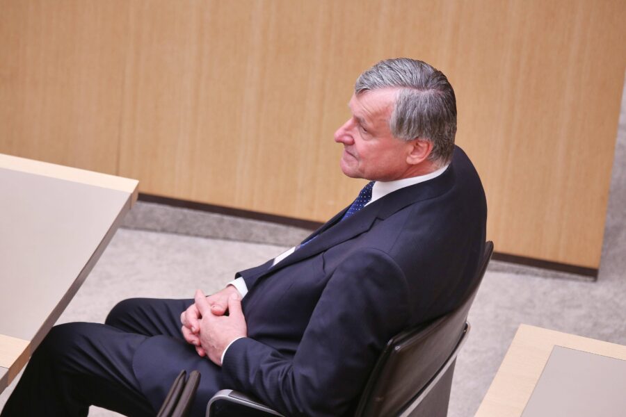 Ein Mann in Anzug sitzt an einem Tisch, Hände gefaltet, Seitenansicht.