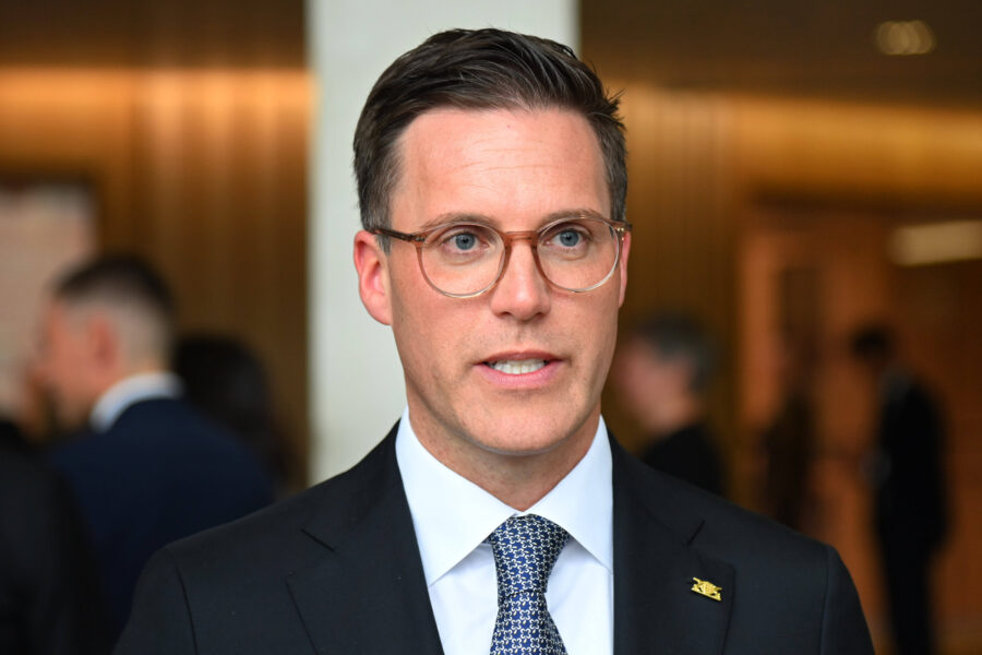 Mann mit Brille und Anzug in einem Innenraum, verschwommene Personen im Hintergrund.