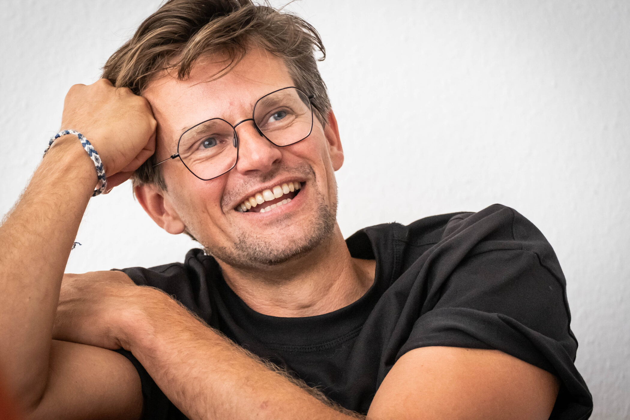 Mann mit Brille lächelt, stützt Kopf mit Hand, trägt schwarzes T-Shirt.