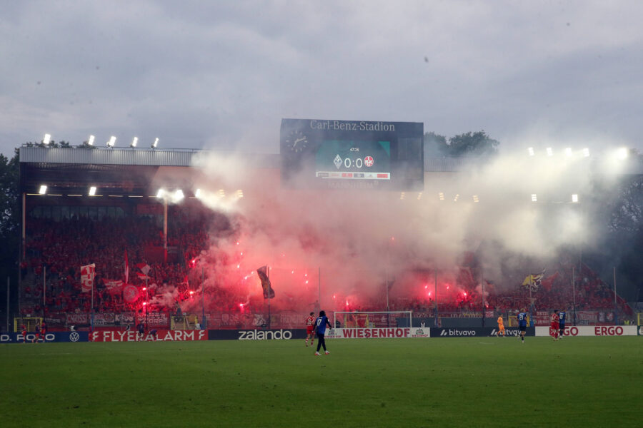 Rauchschwaden im Stadion, rote Fackeln, Zuschauer mit Fahnen auf den Rängen. Spielstand 0:0.