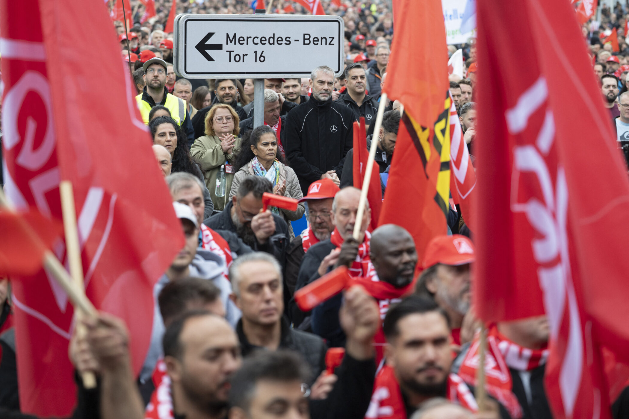 Menschenmenge mit roten Fahnen, Schild: „Mercedes-Benz Tor 16“.