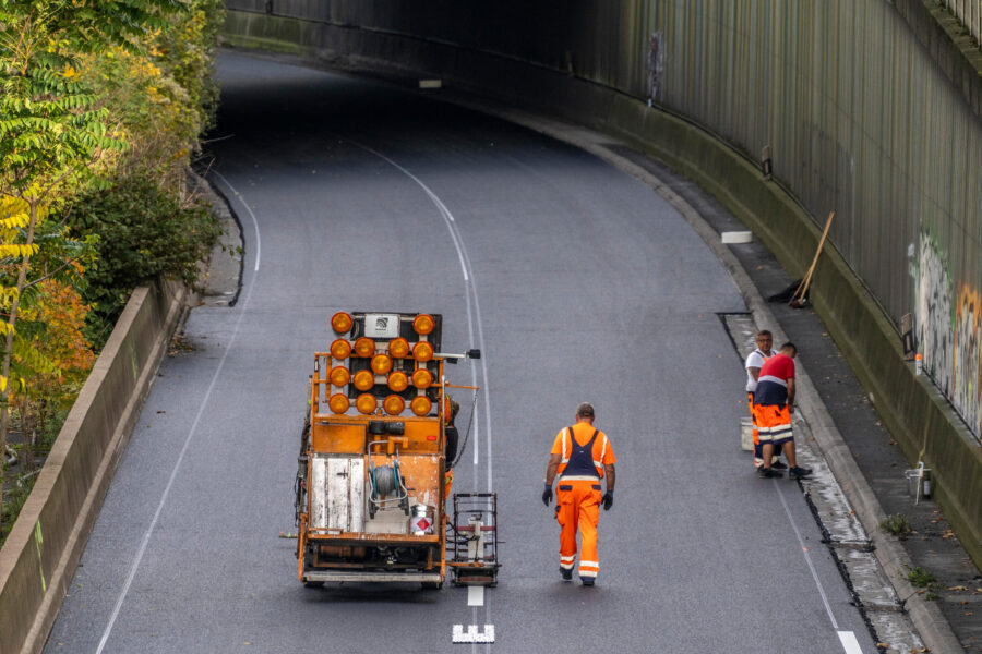 Drei Arbeiter in orangefarbener Kleidung auf einer Straße mit Fahrzeug und Maschinen.