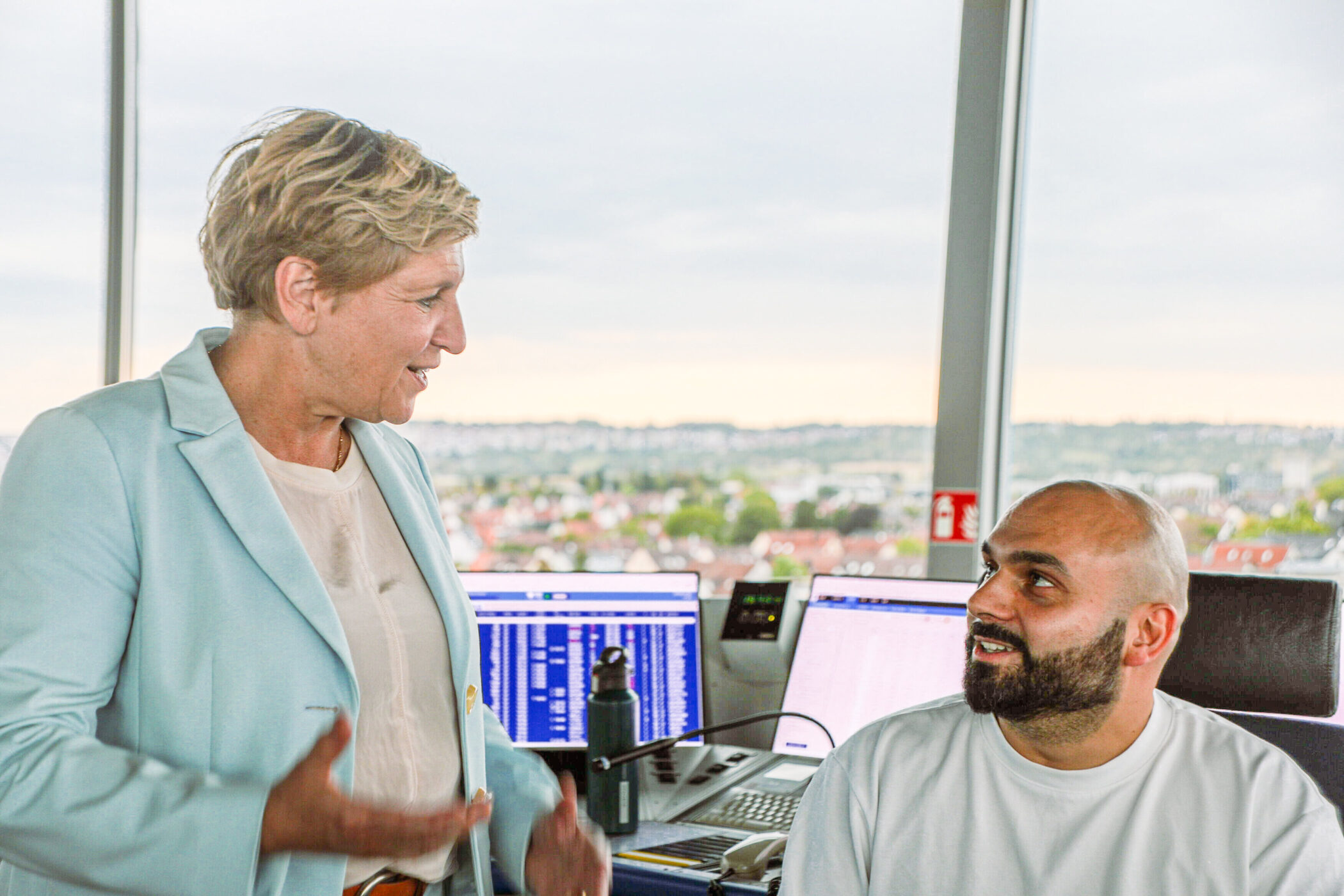 Zwei Personen sprechen in einem Büro mit Computern im Hintergrund.