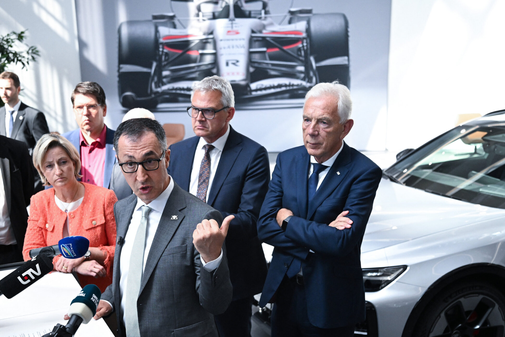 Sechs Personen in Anzug neben einem Auto, Rennwagenbild im Hintergrund.