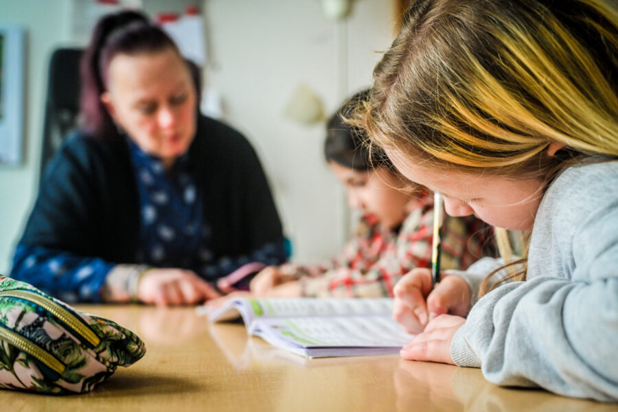 Drei Personen am Tisch, zwei Kinder schreiben in Hefte, erwachsene Person im Hintergrund.