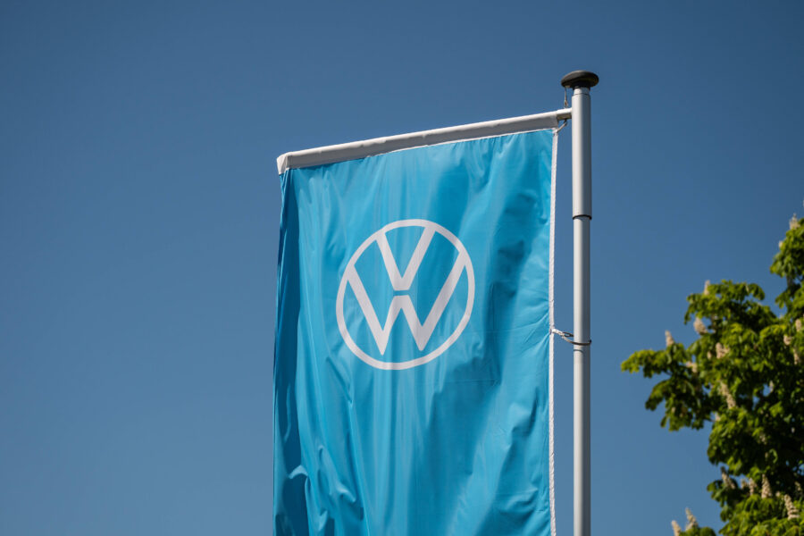 Blaue Fahne mit weißem VW-Logo, klarer Himmel, Baum im Hintergrund.