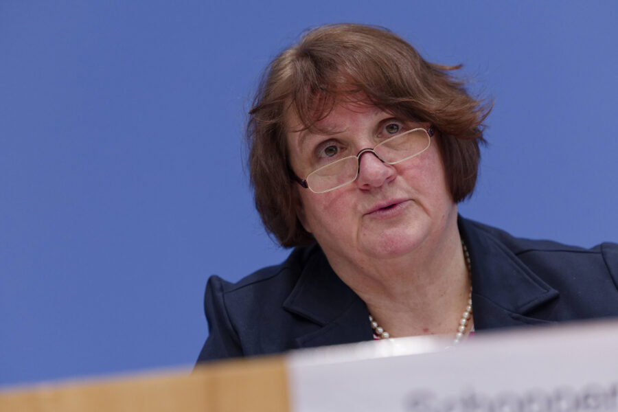 Frau mit Brille und braunem Haar spricht vor blauem Hintergrund.
