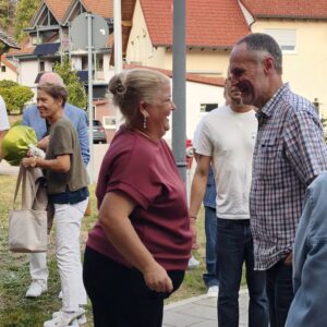 Mehrere Menschen unterhalten sich draußen vor Häusern. Eine Person hält Blumen.