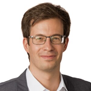 Ein Mann mit Brille und Anzug lächelt vor einem weißen Hintergrund.