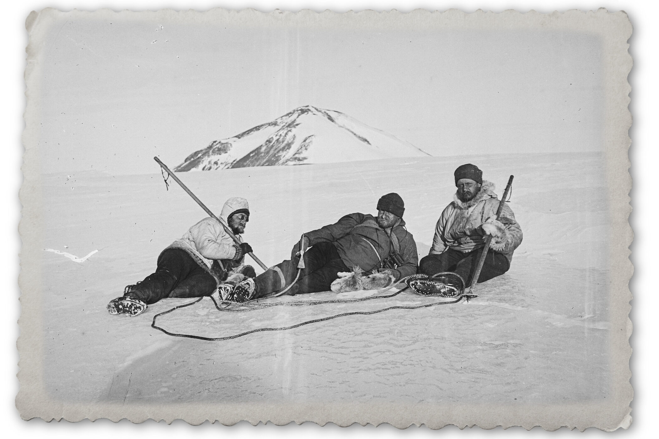 Drei Menschen sitzen auf einem schneebedeckten Feld vor einem Berg.