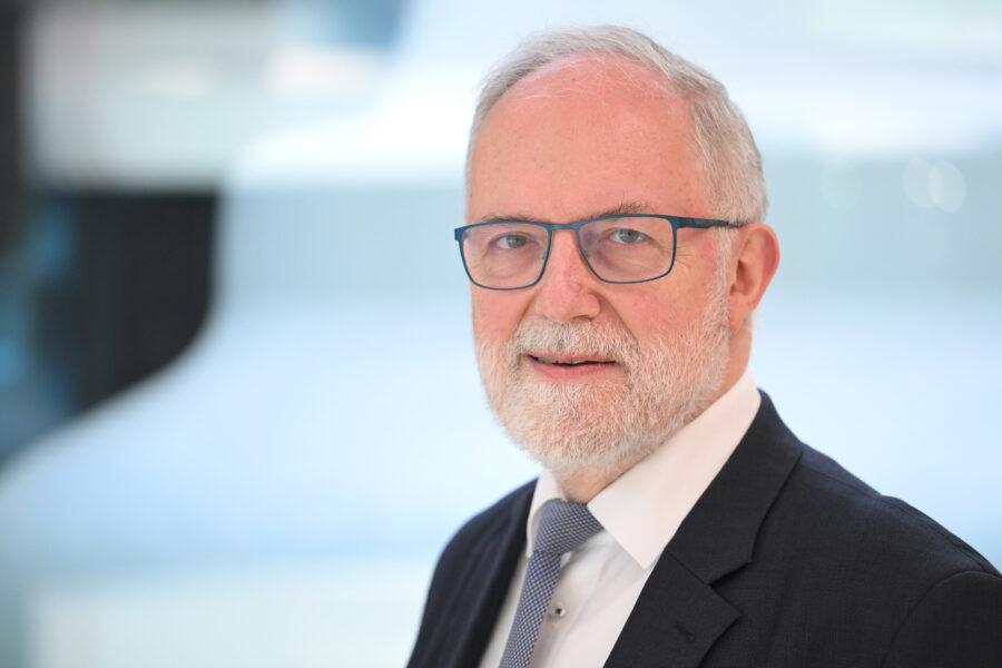 Älterer Mann mit Bart und Brille, trägt Anzug und Krawatte, neutraler Hintergrund.