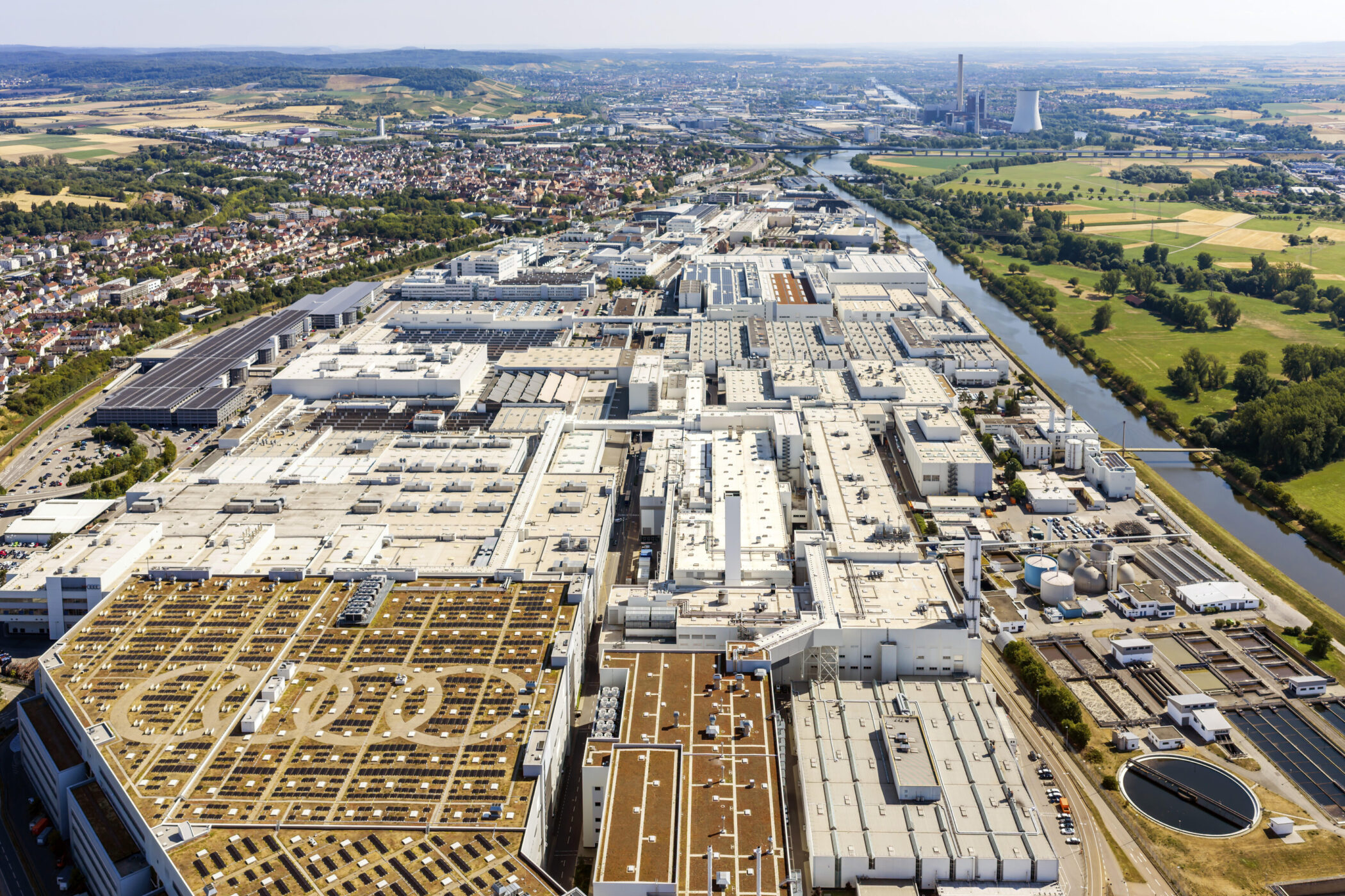 Luftaufnahme großer Industriekomplex mit begrünten Dächern, im Hintergrund Fluss und Stadt.